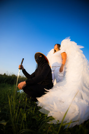 Handsome samurai and angel against blue sky. A masked samurai kneels on a grassy field holding a katana next to a man with large white wings resembling an angel under a clear blue sky.の写真素材