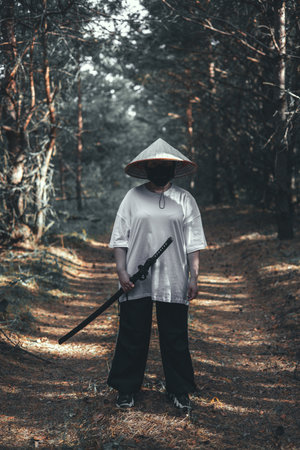 A mysterious warrior in a black mask and Asian hat holds a glittering katana sword in the forest....