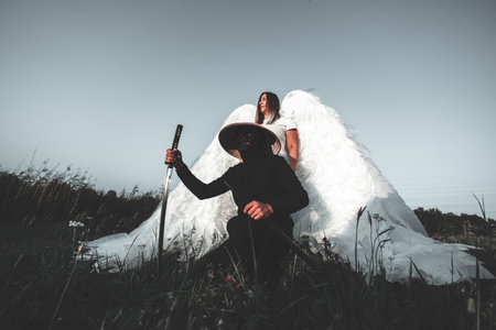 Samurai in black kneeling with katana in twilight, angel behind, mix of cultures and spirituality in serene meadow. Strong warrior and beautiful angel in twilight meadow.の写真素材