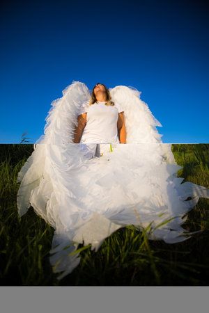A female angel stands against a blue sky. A woman with large white angel wings stands in a grassy field against a blue sky, creating a serene and ethereal scene. Beautiful angel in a field.の写真素材