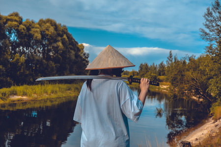 A female samurai stands by a river, holding a katana and looking into the distance on a sunny day. A samurai with a katana on his shoulder contemplates while looking at the calm surface of the river.の写真素材