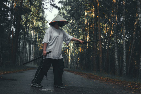 A martial artist in a conical Asian hat practices fighting techniques with a katana sword on a road in the forest. Katana training in the forest against the backdrop of autumn trees.の写真素材