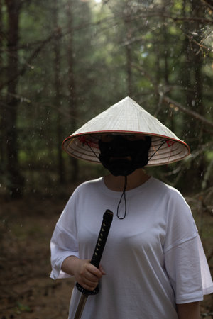 A person in a white shirt and a conical Asian hat holds a katana sword while standing in a forest. Rain falls on the trees in the background.の写真素材