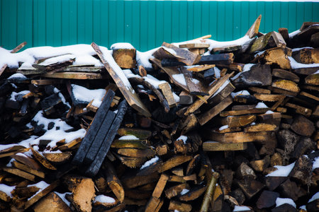 A pile of charred boards after a fire. Snow covering a pile of charred boards near a green fence creates a striking contrast, highlighting the effects of a devastating fire during the winter season.の写真素材