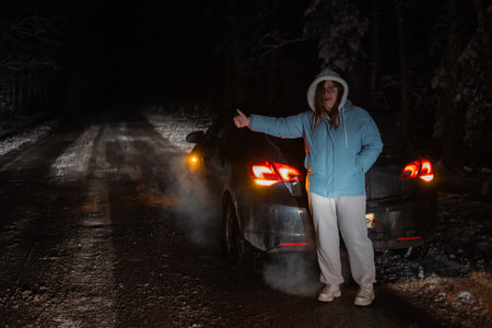 Woman stands beside her broken-down car on a dark, snowy road through a winter forest, signaling for assistance. The scene captures the isolation and urgency of a nighttime roadside emergency.の写真素材