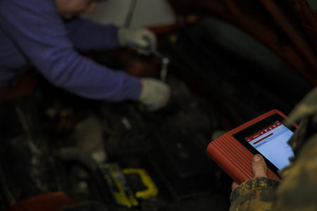 Auto mechanics collaborating in a garage, using electronic diagnostic software on a tablet to analyze a vehicle engine, while another mechanic skillfully applying a wrench for repairs.の写真素材
