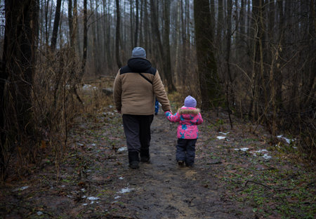 A father and daughter dressed in warm winter clothes enjoy a peaceful walk hand in hand along a muddy forest path, creating a touching moment of family togetherness. Father and daughter walking.の写真素材