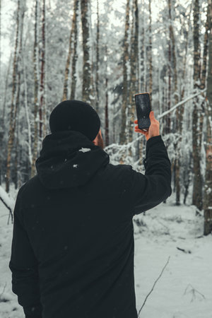 Photographer dressed in black winter clothing is capturing images of a serene snowy birch forest, smartphone held in his right hand, surrounded by tranquil nature and ice-covered trees.の写真素材