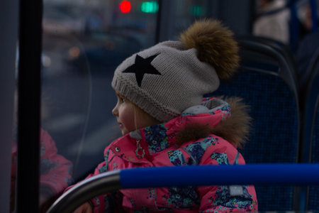 A child rides a bus. A small child in a warm winter outfit looks out the bus window, enchanted by the cityscape. A cozy hat and a colorful jacket emphasize the cold season outside.の写真素材