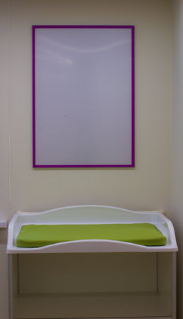 White baby changing table with green mattress and empty purple frame hanging on the wall in a nursery, representing childcare and baby care facilities.の写真素材