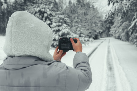 Photographer dressed in cozy winter attire capturing images of a snowy road winding through a serene forest during winter, using a digital camera to document the tranquil landscape.の写真素材
