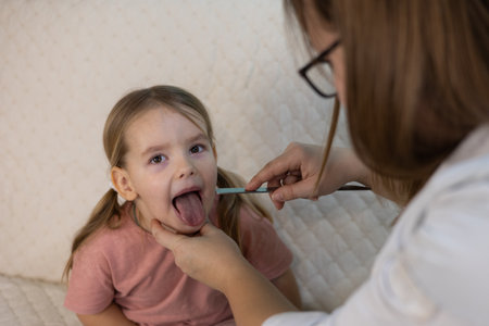 Female pediatrician examines throat of sick little girl with spatula during home visit. Doctor...