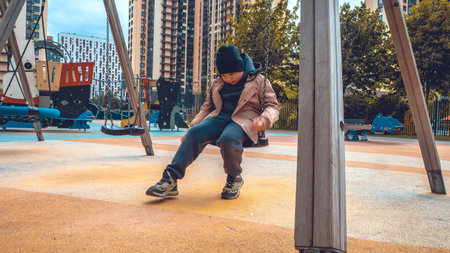 Sad child wearing a beanie and a beige jacket sitting alone on a swing in a deserted playground located in a residential area, conveying a sense of loneliness and abandonment.の写真素材