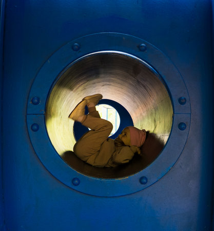 A girl dressed in casual clothes and a pink cap happily slides through a blue tunnel on a playground, enjoying a fun day in the warm sun. Child playing in tunnels on a playground.の写真素材