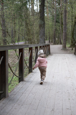 A small child happily runs along a wooden bridge in a quiet forest park, enjoying the beauty of spring, exploring nature and enjoying carefree moments of childhood. A small child runs along a bridge.の写真素材