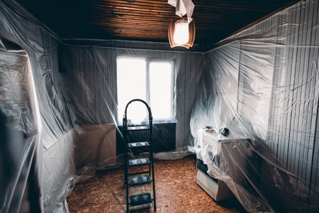 Room undergoing renovation, featuring a ladder in the center and furniture covered with plastic sheets. The wooden ceiling and bright window create a contrast with the protective coverings.の写真素材