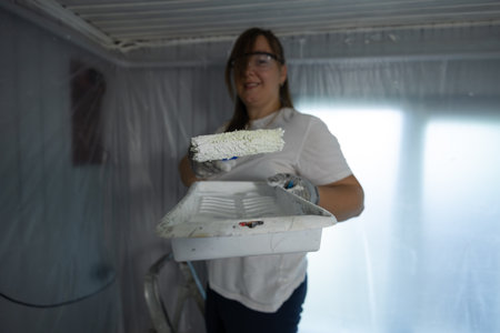 A woman in protective gear paints a room with a roller. Plastic sheeting covers the walls, providing a clean finish. Bright natural light enhances the atmosphere of the workspace.の写真素材