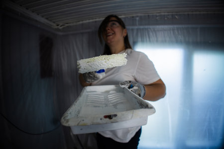 Woman painting a room with a roller brush, wearing protective gear. The space is covered in plastic sheeting, indicating a renovation or home improvement project in progress.の写真素材