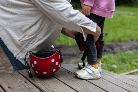 A woman helps her little daughter put on protective gear. The child is preparing to ride a scooter or skateboard. The red helmet is nearby, while mom helps the child wear protective equipment.の写真素材