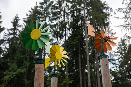 Colourful metal pinwheels spin in the wind, mounted on wooden poles against a backdrop of tall evergreen trees, creating a playful and whimsical scene in a natural setting.の写真素材