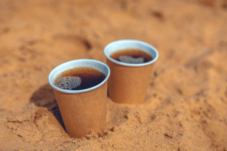 Two disposable coffee cups filled with frothy coffee resting in warm sand, creating a delightful contrast against the earthy tones of the beach environment.の写真素材