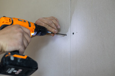 Person expertly uses an orange power drill to secure a screw into drywall, showcasing precision and skill in construction work. The focus is on the hands and the drill in action.の写真素材