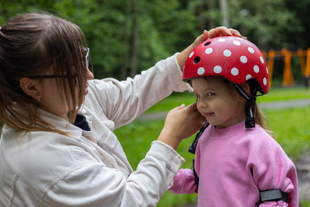 A young mother places a red helmet with white polka dots on her smiling daughters head before riding a scooter in the park on a sunny summer day. Child safety while riding a scooter.の写真素材