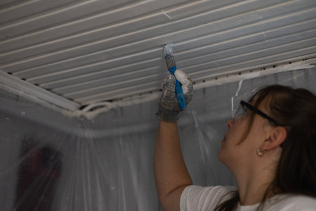 A female painter wearing safety glasses and gloves applies white paint to a ceiling with a brush while working on a home renovation project. The woman carefully paints the ceiling with white paint.の写真素材