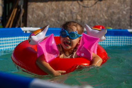 Happy little girl swimming and playing in a home pool, wearing inflatable armbands and a bright red swim ring, enjoying carefree moments during summer holidays under the sunny sky.の写真素材
