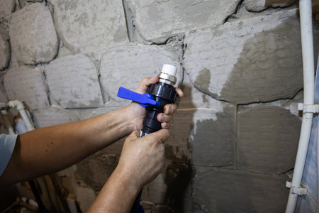 Human hands are connecting a black plastic plumbing valve with a blue handle to a flexible pipe, installing a new section of a water distribution system against a rough concrete wall.の写真素材
