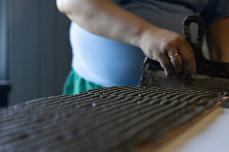 A worker applies adhesive to a tile with a notched trowel. The adhesive is evenly distributed across the rough surface of the tile. A close-up shows the texture and skill of the hands.の写真素材