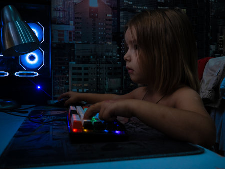 Young child focusing intently on a computer screen, hands on a colorful backlit keyboard and mouse,...