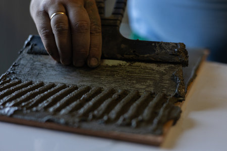 A persons hand applies mortar with a notched trowel to the back of a tile. A close-up of textured adhesive and the skillful work of tiling during a home renovation or flooring installation.の写真素材