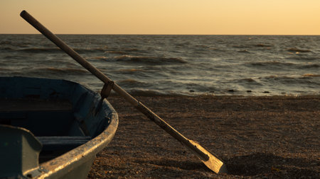 An old wooden oar balances on a small blue rowboat resting on the beach during the calm golden hour. Against the backdrop of calm waves, the old boat creates a serene and tranquil coastal landscape.の写真素材