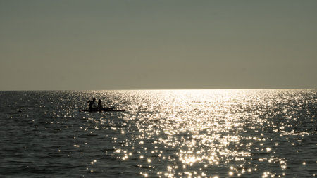 Two people paddleboarding on a calm ocean, silhouetted against the setting sun reflecting brightly and sparkling on the water surface, creating a peaceful and serene scene.の写真素材