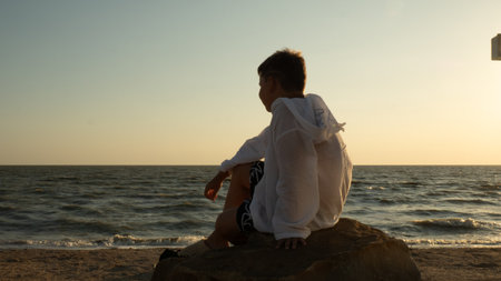 A boy sits on a coastal rock during golden hour, quietly and thoughtfully gazing at the ocean horizon. A teenager sits on the seashore at sunset, serene and peaceful, pensively gazing at the waves.の写真素材