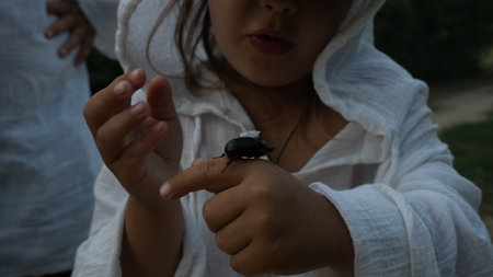 A child wearing a white hooded shirt carefully touches a large beetle crawling on his arm. The child...