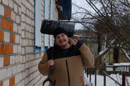 A man smiles at the camera holding a snow shovel over his shoulder clearing the yard in front of a...