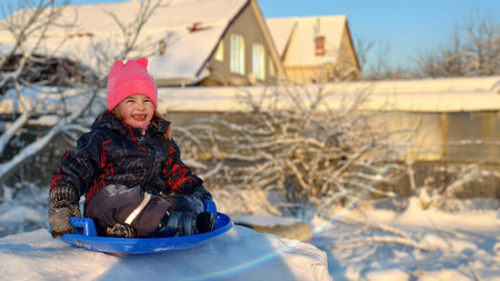 Cheerful young child wearing a pink hat and winter clothing, sitting on a blue sled, laughing and playing on a snowy outdoor day with houses and bare trees in background.の写真素材