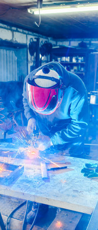 Vertical. A welder wearing a protective mask performs arc welding of metal parts, generating bright...