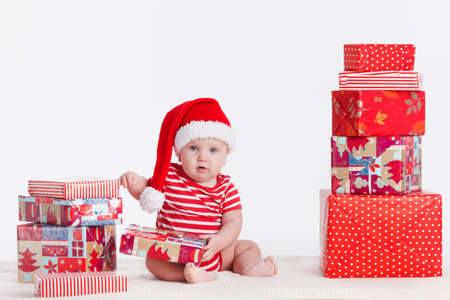 Adorable child is sitting on floor, wearing red Christmas cap, ired balls and presents around  isolated on white backgroundの写真素材