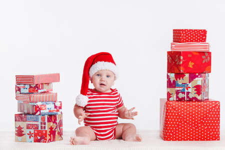 Adorable child is sitting on floor, wearing red Christmas cap, ired balls and presents around ...