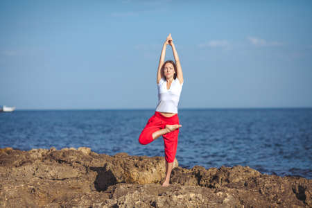Beautiful positive blond girl clothing in white sit at the seaside on the rock and meditating in yoga poseの写真素材