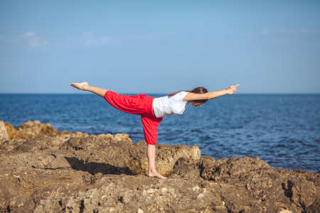 Beautiful positive blond girl clothing in white sit at the seaside on the rock and meditating in yoga poseの写真素材