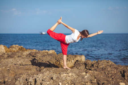 Beautiful positive blond girl clothing in white sit at the seaside on the rock and meditating in yoga poseの写真素材
