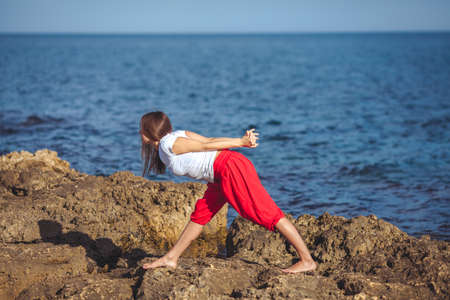 Beautiful positive blond girl clothing in white sit at the seaside on the rock and meditating in yoga poseの写真素材