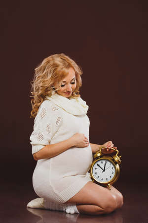 portrait of pregnant woman with clock in her hands sitting on brown backgroundの写真素材