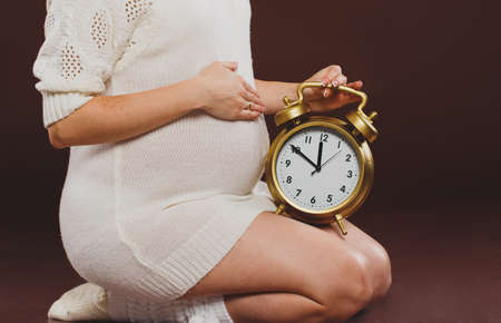 portrait of pregnant woman with clock in her hands sitting on brown backgroundの写真素材