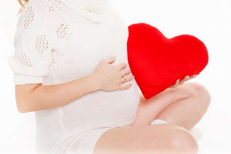 Pregnant beautiful woman holding red heart pillow in her hands isolated on white background