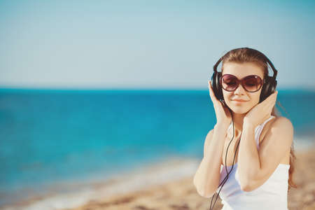 Beautiful woman listening to music against blue sky in headphones on the seashoreの写真素材